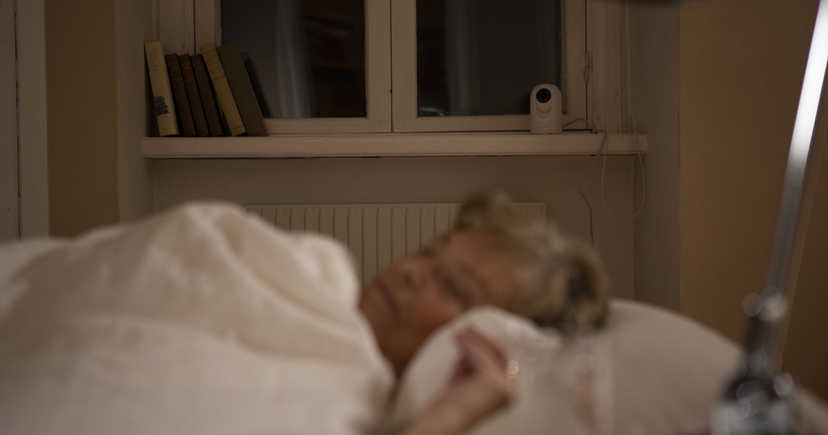 En äldre kvinna ligger i sin säng. I bakgrunden syns en kamera som finns till för att kvinnan ska få tillsyn digitalt.